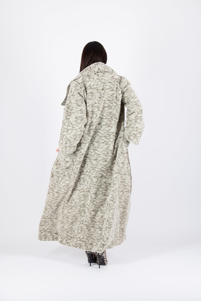 May include: A long, textured coat in shades of beige and gray, with a subtle pattern. The coat has a wide collar and long sleeves. The hemline reaches the ankles. The person is wearing patterned high heels.