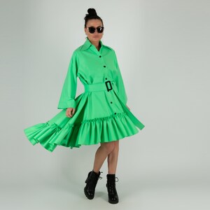 May include: A bright green dress with long sleeves, a belted waist, and a ruffled skirt. The dress has a button-down front and is paired with black boots. The model is wearing sunglasses.