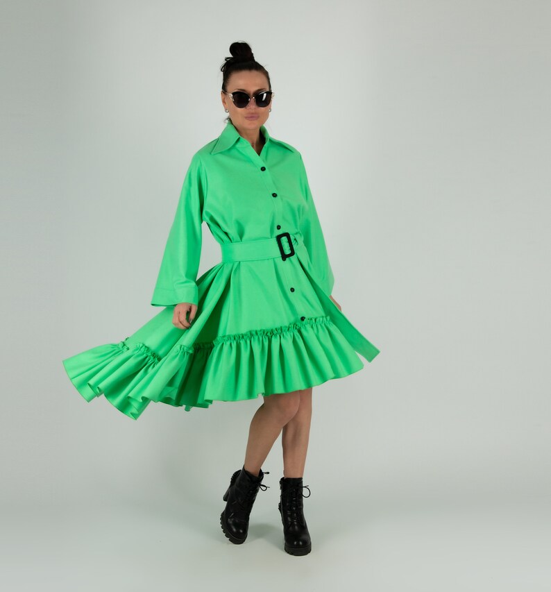 May include: A bright green dress with long sleeves, a belted waist, and a ruffled skirt. The dress has a button-down front and is paired with black boots. The model is wearing sunglasses.