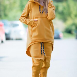 May include: A mustard yellow hooded sweatshirt and matching sweatpants with a black drawstring. The sweatshirt has a large kangaroo pocket and a high-low hemline.