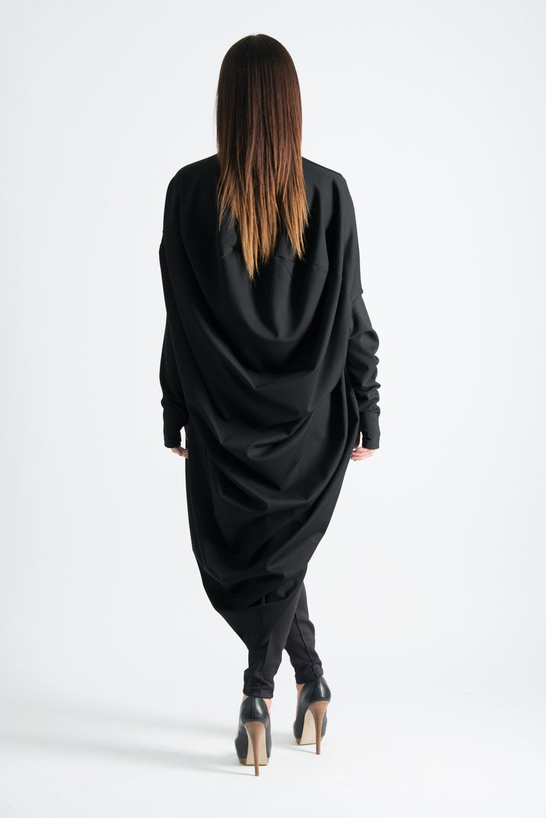 May include: A black long-sleeved dress with a draped design. The dress is fitted at the waist and flows out to the hem. The model is wearing black heels.