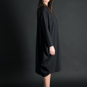 May include: A black long-sleeved dress with a loose fit and a slightly gathered skirt. The dress is worn by a person with long dark hair and sunglasses. The person is standing on a black background.