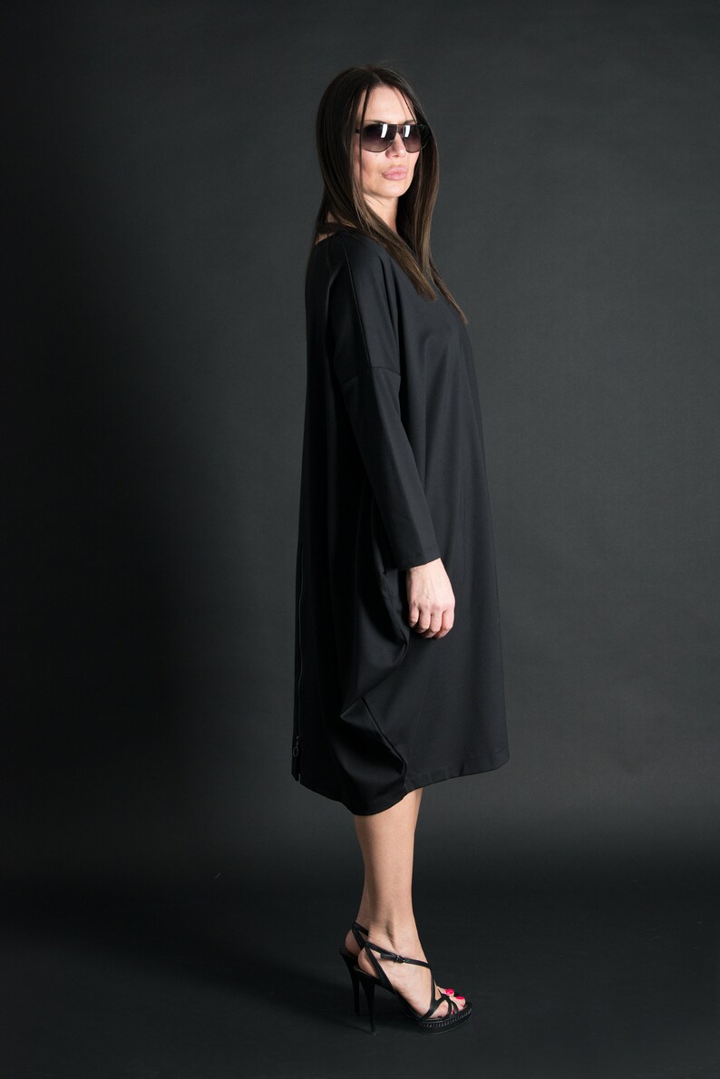 May include: A black long-sleeved dress with a loose fit and a slightly gathered skirt. The dress is worn by a person with long dark hair and sunglasses. The person is standing on a black background.