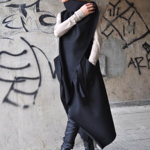 May include: A long black asymmetrical vest with a high collar and a tie detail. The vest is worn over a beige long-sleeved shirt. The person is wearing black pants and black boots.