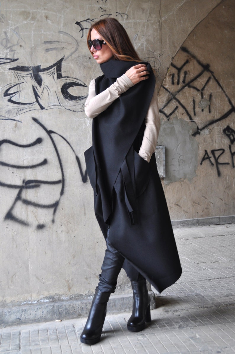 May include: A long black asymmetrical vest with a high collar and a tie detail. The vest is worn over a beige long-sleeved shirt. The person is wearing black pants and black boots.
