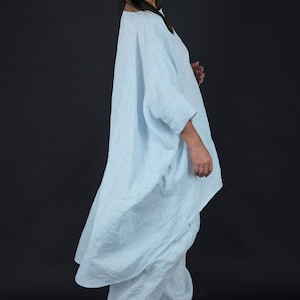 May include: A light blue linen tunic with a loose, flowing fit. The tunic has a wide, round neckline and long sleeves. The model is wearing the tunic with white linen pants and black heels.