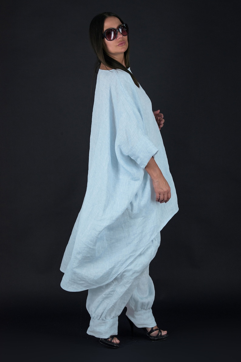 May include: A light blue linen tunic with a loose, flowing fit. The tunic has a wide, round neckline and long sleeves. The model is wearing the tunic with white linen pants and black heels.