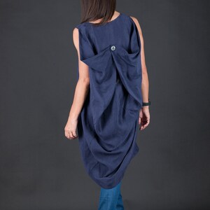 May include: A navy blue sleeveless dress with a gathered back detail and a single button closure. The dress is worn with blue jeans and brown heeled shoes.