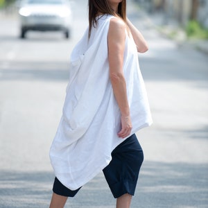May include: A woman wearing a white linen tunic top with a loose, flowing silhouette. She is also wearing dark blue cropped pants and white heeled sandals.
