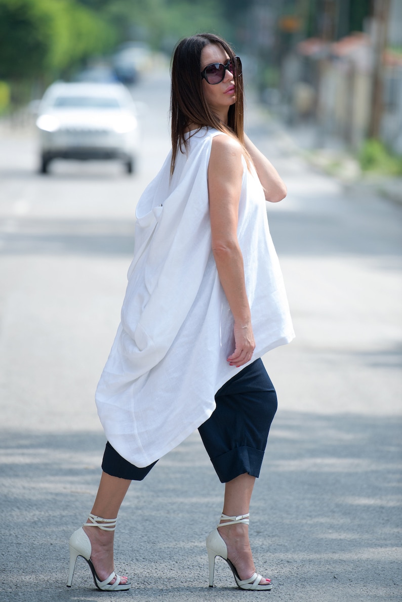 May include: A woman wearing a white linen tunic top with a loose, flowing silhouette. She is also wearing dark blue cropped pants and white heeled sandals.