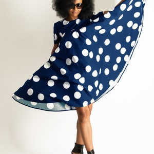 May include: A navy blue dress with large white polka dots. The dress has short sleeves and is being held up to show its flowing design. The person in the image is wearing black high-heeled sandals and sunglasses.