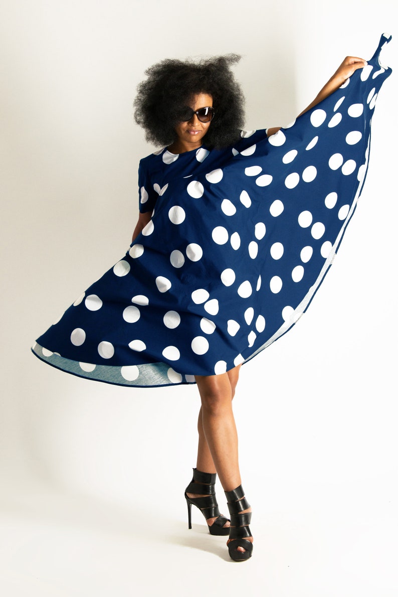 May include: A navy blue dress with large white polka dots. The dress has short sleeves and is being held up to show its flowing design. The person in the image is wearing black high-heeled sandals and sunglasses.