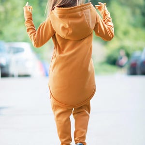 May include: A mustard yellow hooded sweatshirt with a long, fitted design. The sweatshirt has a zipper closure and a drawstring hood. The model is wearing the sweatshirt with matching pants.