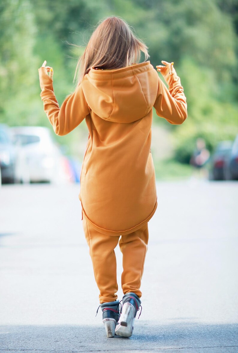 May include: A mustard yellow hooded sweatshirt with a long, fitted design. The sweatshirt has a zipper closure and a drawstring hood. The model is wearing the sweatshirt with matching pants.