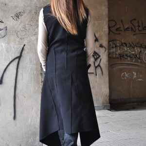 May include: A long black vest with a high neckline and a asymmetrical hemline. The vest is made of a thick fabric and has a raw edge detail. The model is wearing the vest over a white long-sleeved shirt and black pants.