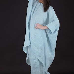 May include: A light blue linen tunic with a long, flowing silhouette. The tunic has a button closure at the neck and a relaxed fit. The model is wearing the tunic with black heels.