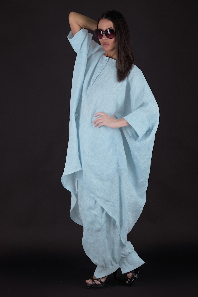 May include: A light blue linen tunic with a long, flowing silhouette. The tunic has a button closure at the neck and a relaxed fit. The model is wearing the tunic with black heels.