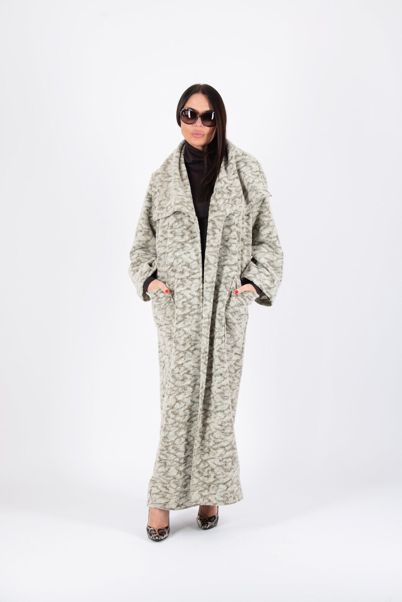 May include: A long, light gray coat with a textured pattern. The coat has large pockets and a wide collar. The person is wearing sunglasses and a black turtleneck. The shoes have a leopard print.