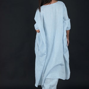 May include: A light blue linen dress with a loose, flowing fit. The dress has a scoop neckline and three-quarter sleeves. The model is wearing the dress with a pair of white linen pants.
