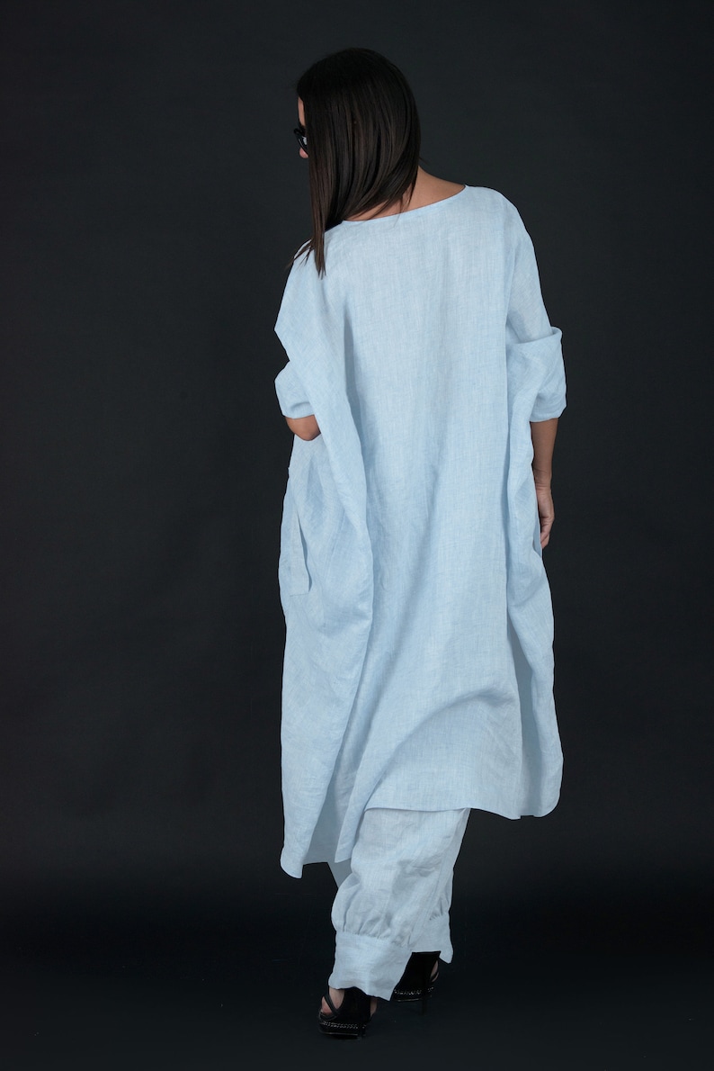 May include: A light blue linen dress with a loose, flowing fit. The dress has a scoop neckline and three-quarter sleeves. The model is wearing the dress with a pair of white linen pants.