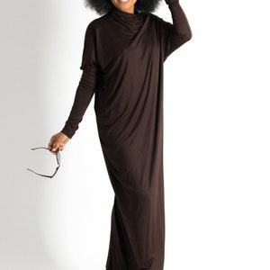 May include: A woman with curly black hair is wearing a long brown maxi dress. She is smiling and holding a pair of sunglasses.
