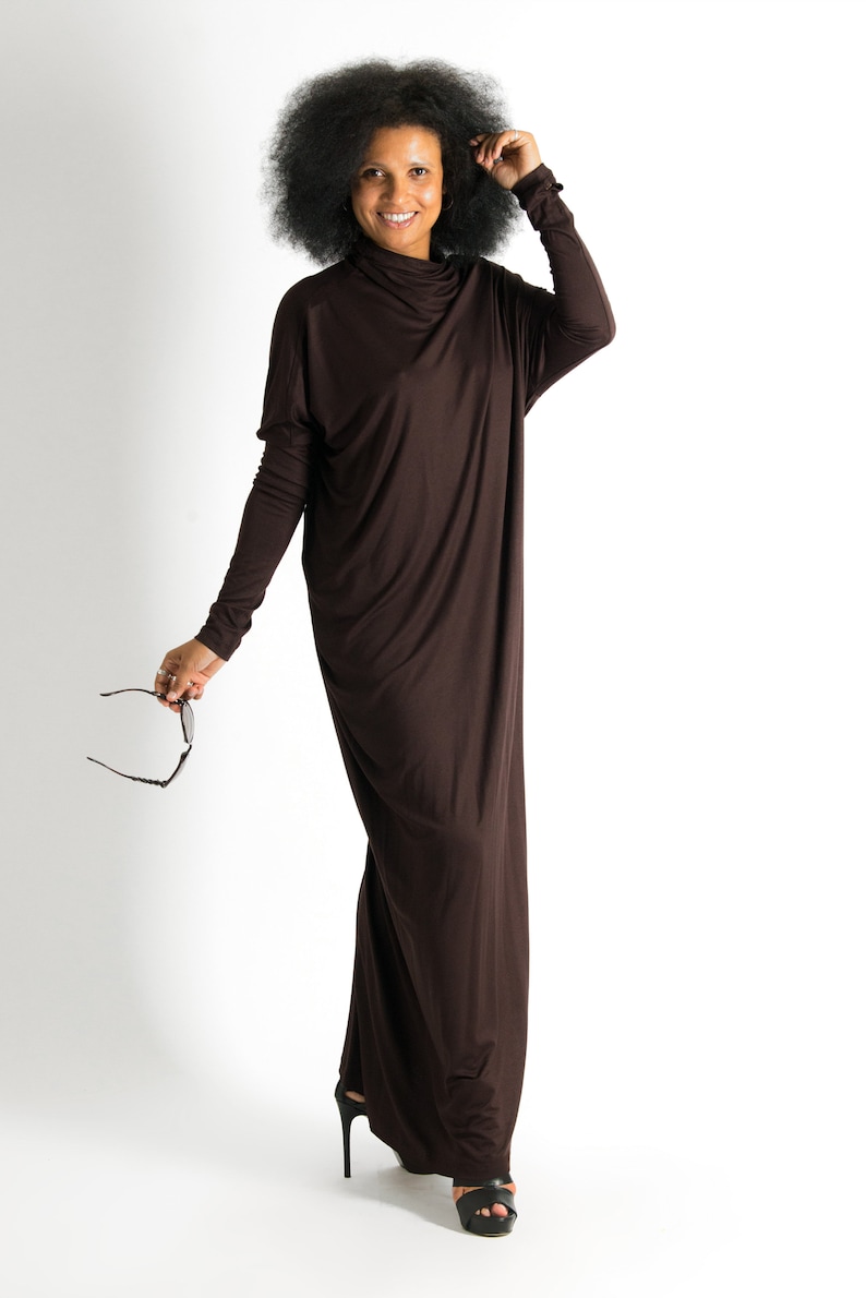 May include: A woman with curly black hair is wearing a long brown maxi dress. She is smiling and holding a pair of sunglasses.