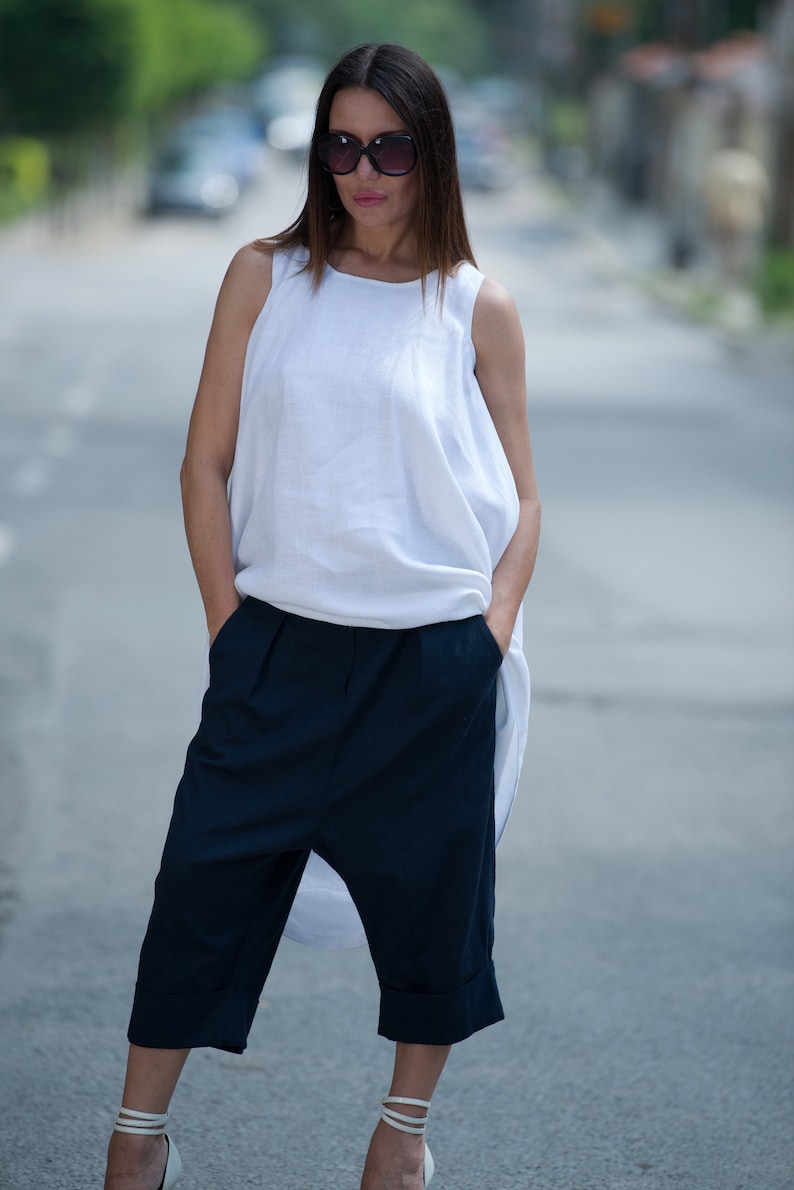 May include: A woman wearing a white linen top with a loose, flowing silhouette and dark blue, cropped, wide-leg pants. She is wearing sunglasses and white heeled sandals.