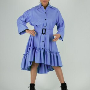 May include: A light blue dress with a button-down front, long sleeves, and a belted waist. The dress features a ruffled hemline and is paired with black boots. The model is wearing sunglasses.