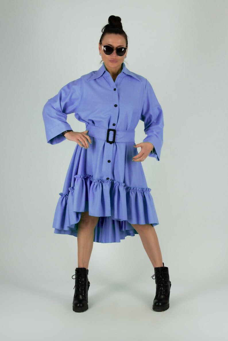 May include: A light blue dress with a button-down front, long sleeves, and a belted waist. The dress features a ruffled hemline and is paired with black boots. The model is wearing sunglasses.
