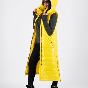 May include: A long yellow puffer vest with a hood. The vest has a zipper closure and a quilted design. The model is wearing black pants and black heeled boots.