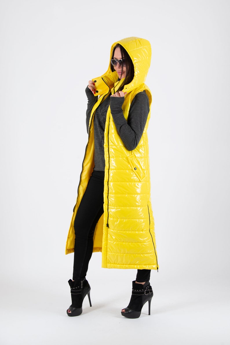 May include: A long yellow puffer vest with a hood. The vest has a zipper closure and a quilted design. The model is wearing black pants and black heeled boots.