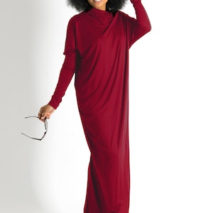 May include: A woman wearing a long, burgundy maxi dress with a cowl neck. She is standing on a white background and is holding a pair of sunglasses.