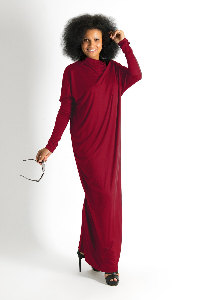 May include: A woman wearing a long, burgundy maxi dress with a cowl neck. She is standing on a white background and is holding a pair of sunglasses.