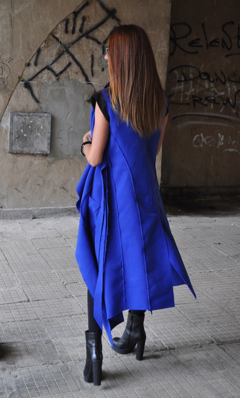 Peut inclure: Une femme portant un long gilet bleu royal au design asym&eacute;trique unique. Le gilet a un bord effiloch&eacute; et est fait d'un tissu &eacute;pais et textur&eacute;. Elle porte des bottes noires &agrave; talons.