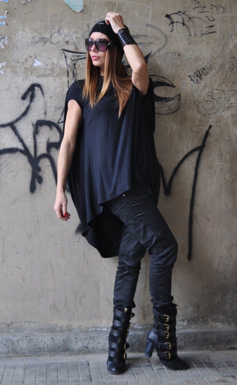 May include: A woman wearing a black oversized shirt, black jeans, and black boots with gold buckles. She is wearing a black beanie and sunglasses.
