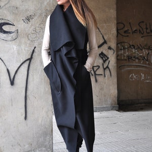May include: A long black sleeveless coat with a draped collar and pockets. The coat is worn over a beige top and black boots.
