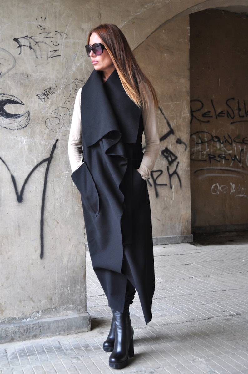 May include: A long black sleeveless coat with a draped collar and pockets. The coat is worn over a beige top and black boots.