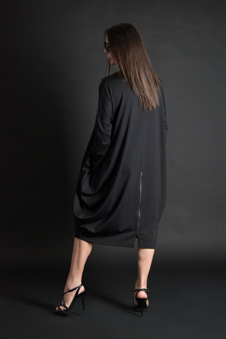 May include: A black long-sleeved dress with a zipper down the back. The dress is loose-fitting and has a gathered look. The model is wearing black high-heeled sandals.