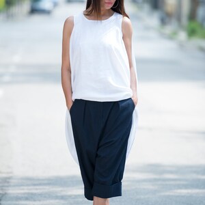 May include: A woman wearing a white sleeveless top and dark blue cropped pants. She is wearing sunglasses and white heeled sandals.