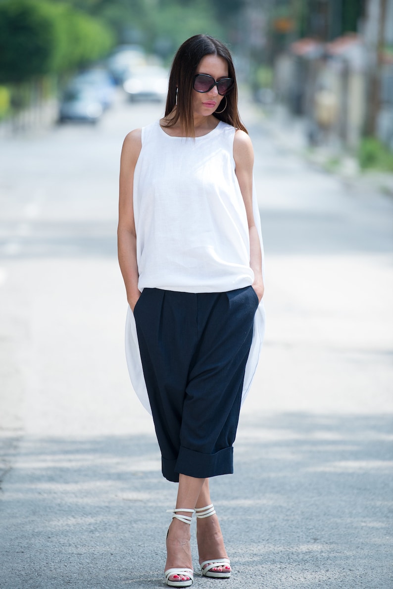 May include: A woman wearing a white sleeveless top and dark blue cropped pants. She is wearing sunglasses and white heeled sandals.