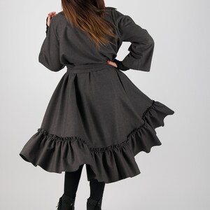 May include: A dark gray coat dress with a flared skirt and ruffled hem. The dress has long sleeves and a matching belt. The model is wearing black leggings and black high-heeled boots with fringe.