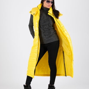 May include: A long yellow puffer vest with a hood. The vest is unzipped and the person wearing it is standing with their legs crossed.