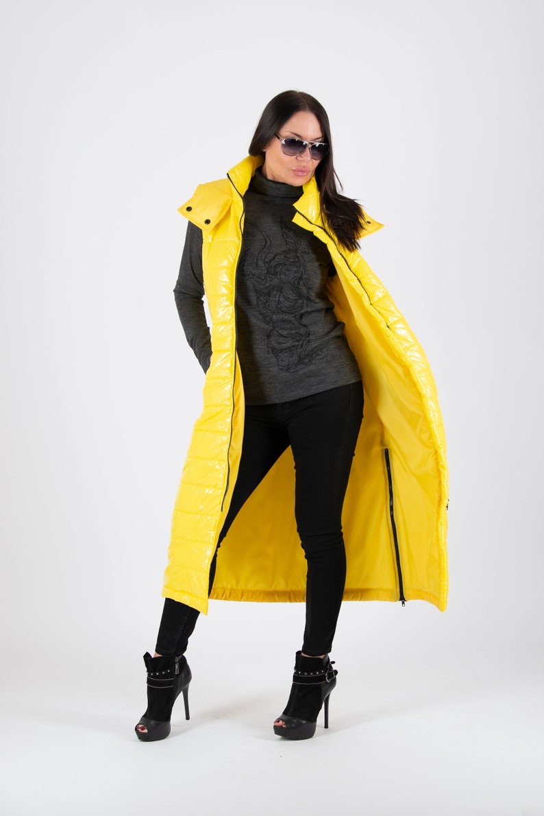 May include: A long yellow puffer vest with a hood. The vest is unzipped and the person wearing it is standing with their legs crossed.