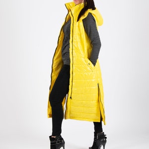 May include: A long yellow puffer vest with a hood and black zipper accents. The vest is worn over a grey long-sleeved shirt and black pants.