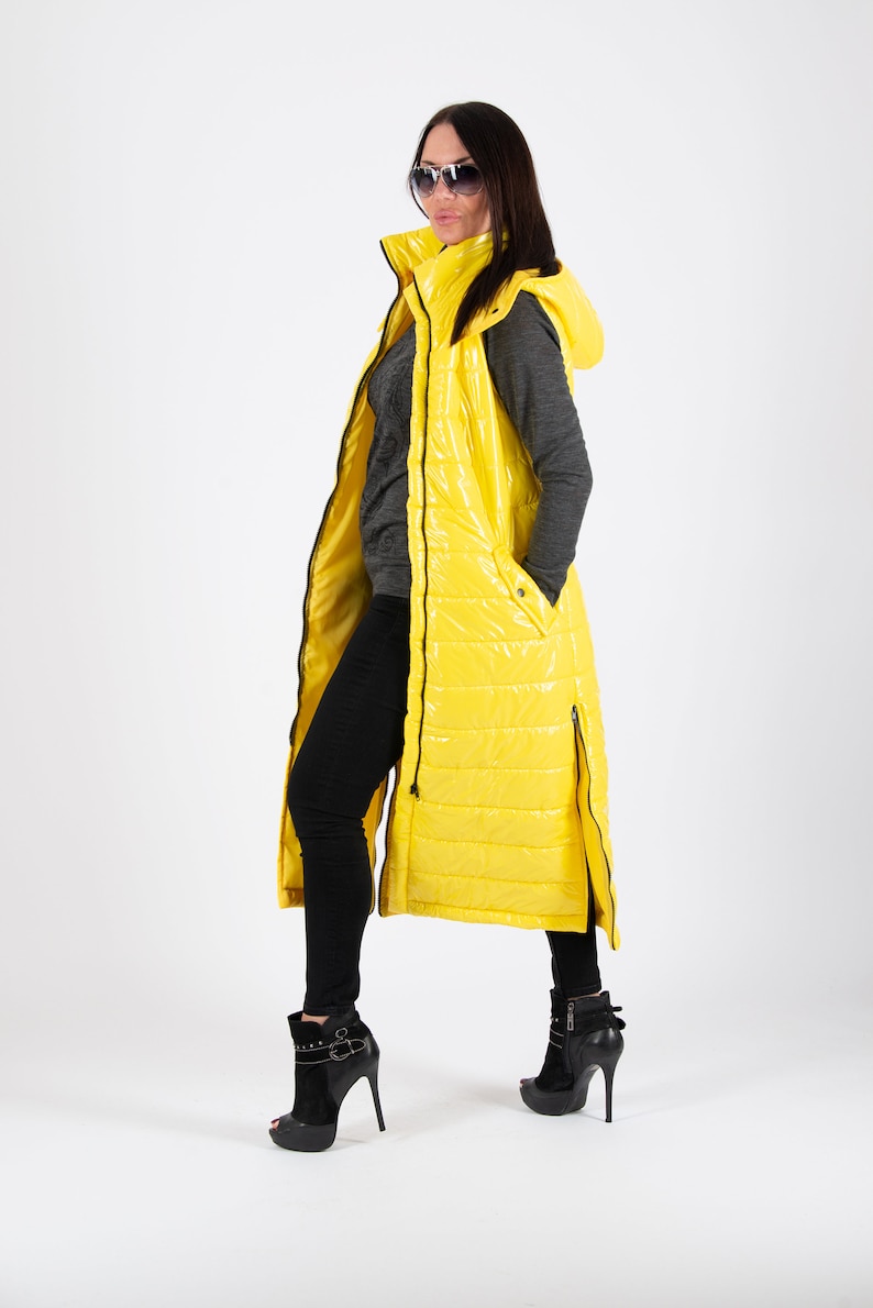 May include: A long yellow puffer vest with a hood and black zipper accents. The vest is worn over a grey long-sleeved shirt and black pants.