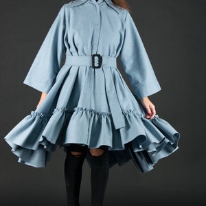 May include: A light blue dress with a belted waist and a flared skirt. The dress features a button-down front, a collar, and long sleeves. The skirt has a ruffled hem. The model is wearing black sunglasses and black heels.