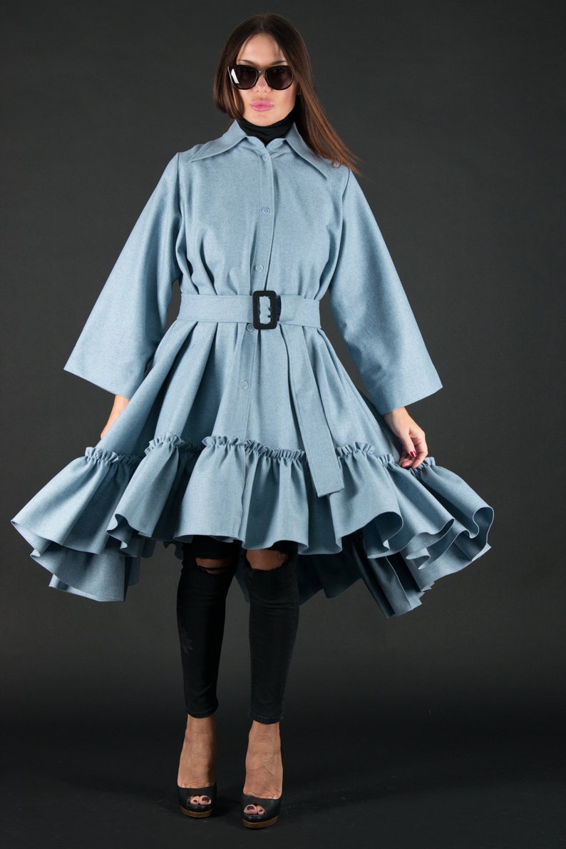 May include: A light blue dress with a belted waist and a flared skirt. The dress features a button-down front, a collar, and long sleeves. The skirt has a ruffled hem. The model is wearing black sunglasses and black heels.