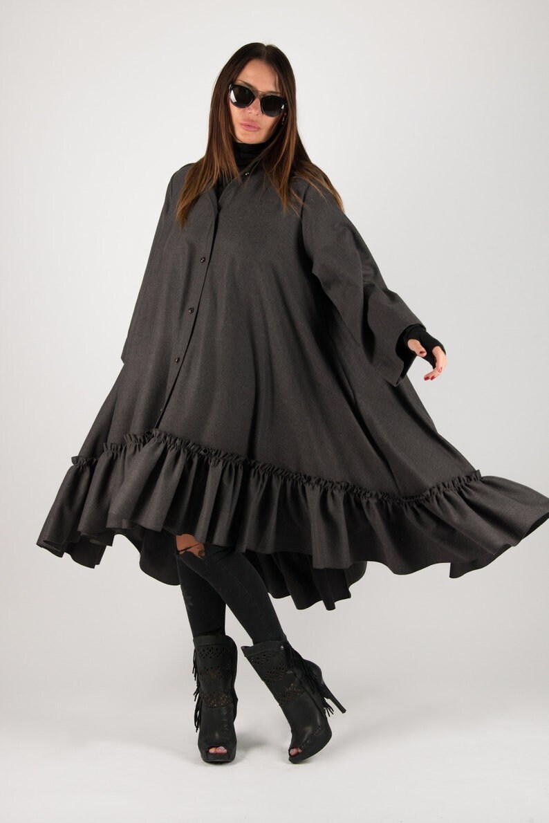 May include: A dark gray, button-down dress with a ruffled hem. The dress has long sleeves and a high-low design. The model is wearing black sunglasses and black boots.