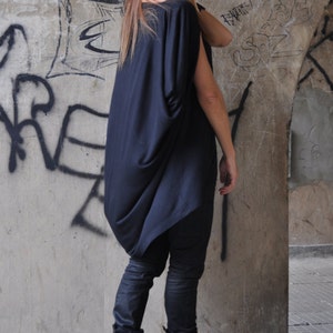 May include: A woman wearing a black, loose-fitting top with a black beanie and black boots. She is standing in front of a concrete wall with graffiti.