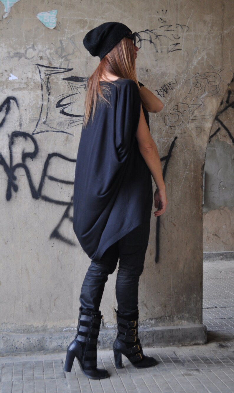 May include: A woman wearing a black, loose-fitting top with a black beanie and black boots. She is standing in front of a concrete wall with graffiti.